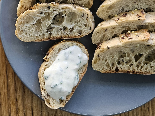 Tranches de pain aux céréales sur une assiette, dont une tartine généreusement garnie de fromage frais aux herbes.